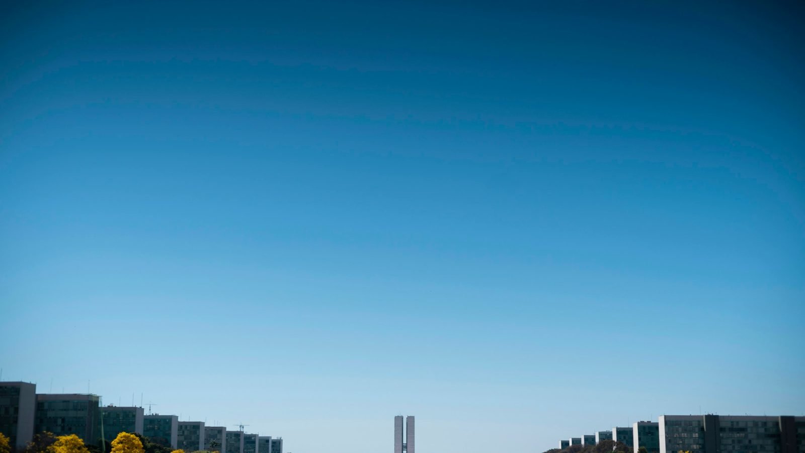 Esplanada dos Ministérios em Brasília, com os blocos dos ministérios dos dois lados e o Congresso Nacional ao fundo sob céu azul